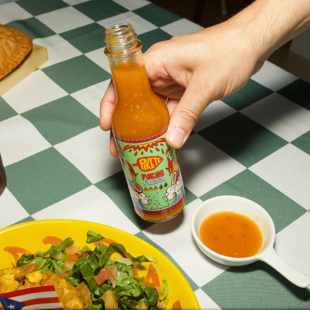 Hand holding a bottle of hot sauce over a plate of food on a checkered tablecloth.