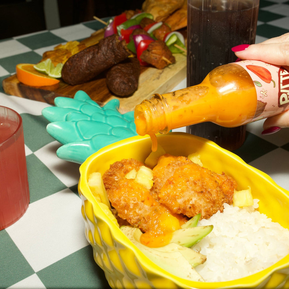 Person pouring hot sauce over fried shrimp  and rice in a pineapple shaped bowl on a checkered tablecloth.