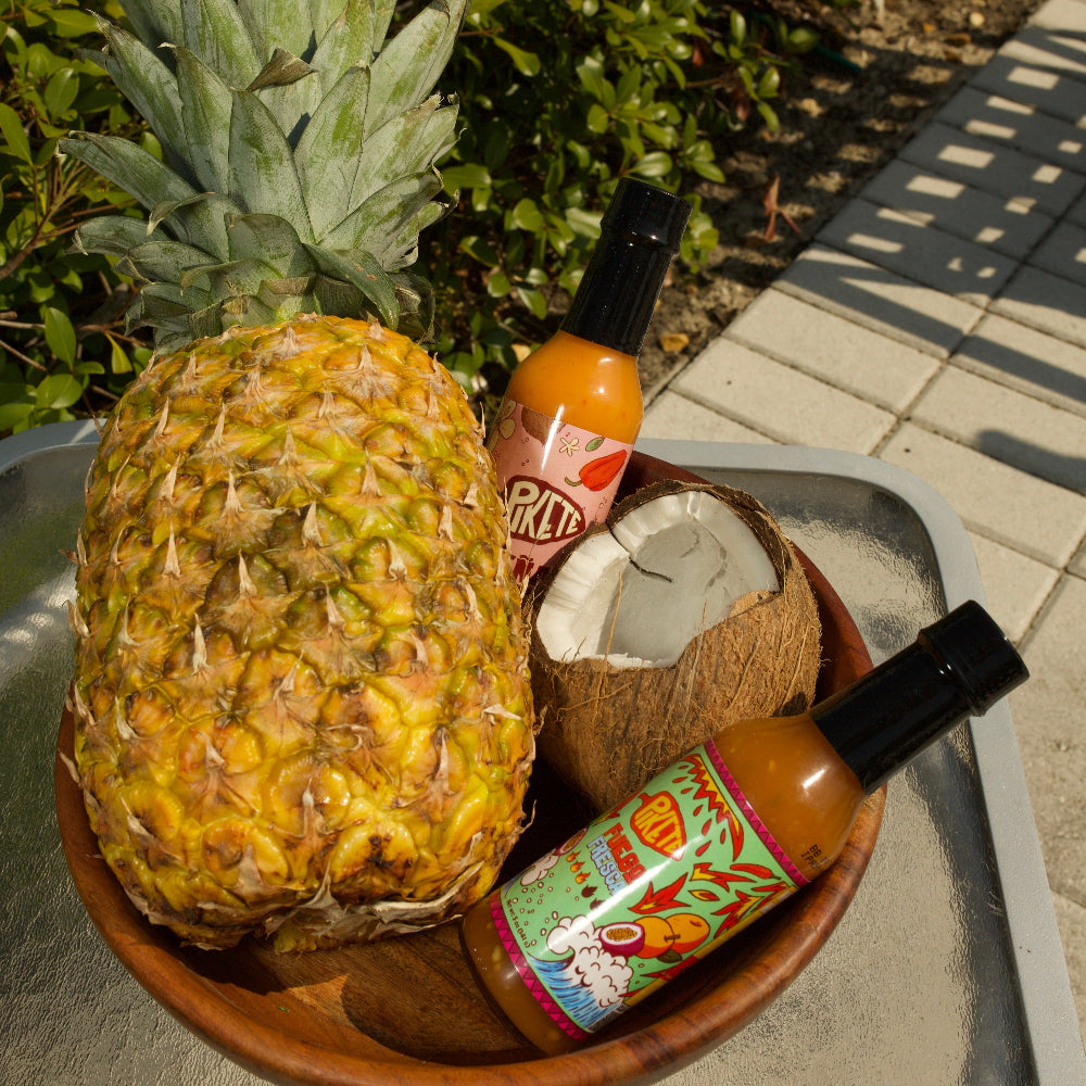 Pineapple, coconut, and hot sauce bottles on a wooden bowl outdoors